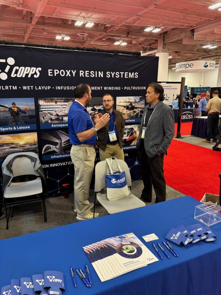 Trade-show booth for COPPS Epoxy Resin Systems with three men talking in front of large banner and promotional materials on a blue table nearby.